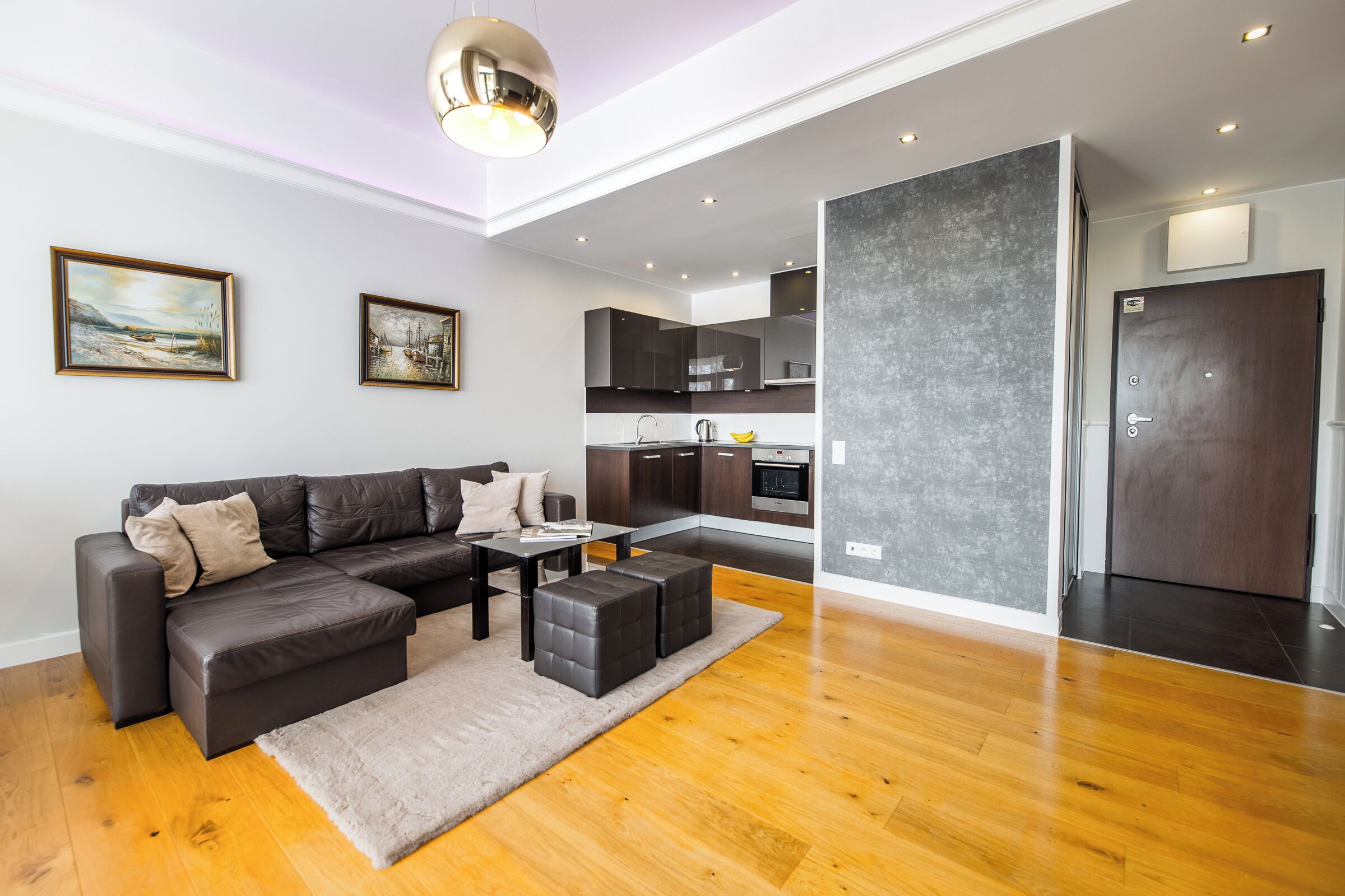 A modern living room with a sectional sofa, a coffee table, and a rug. There is a kitchen area with dark cabinets and a refrigerator.