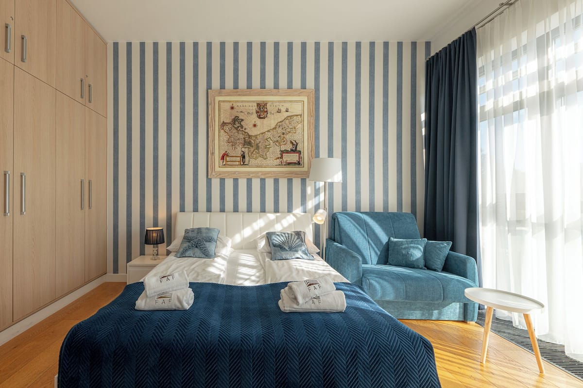 Una habitación de hotel con una cama, un sillón azul, un mapa en la pared y una ventana con cortinas.