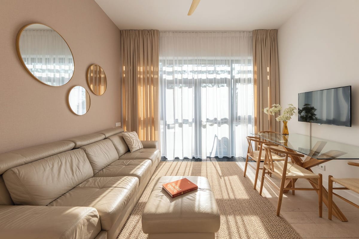 Una sala de estar moderna con un sofá color beige, una mesa de comedor de vidrio con sillas, un televisor montado en la pared y un ventanal con cortinas translúcidas.