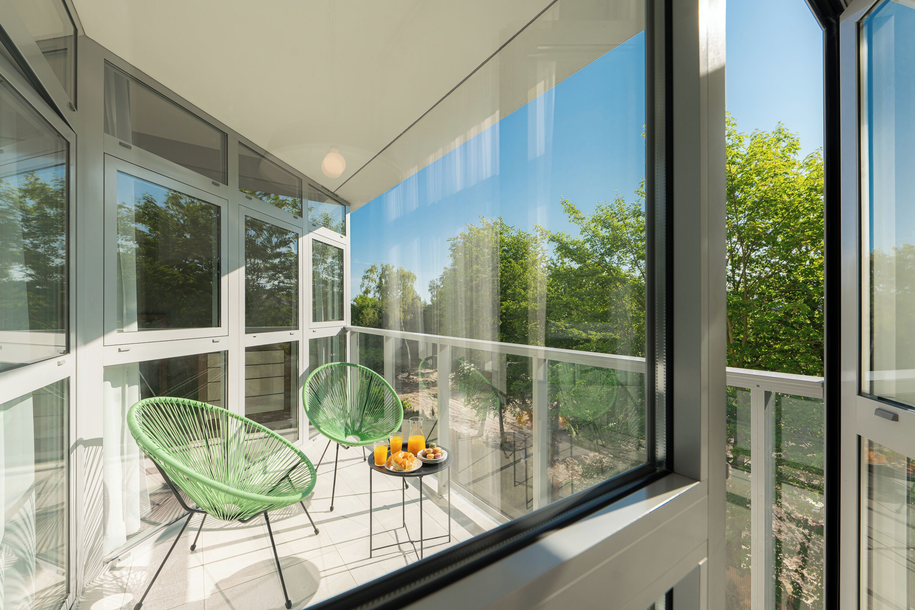 A balcony with glass walls, two green chairs, a small table with a plate of food and a glass of orange juice.