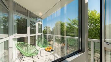 A balcony with glass walls, two green chairs, a small table with a plate of food and a glass of orange juice.