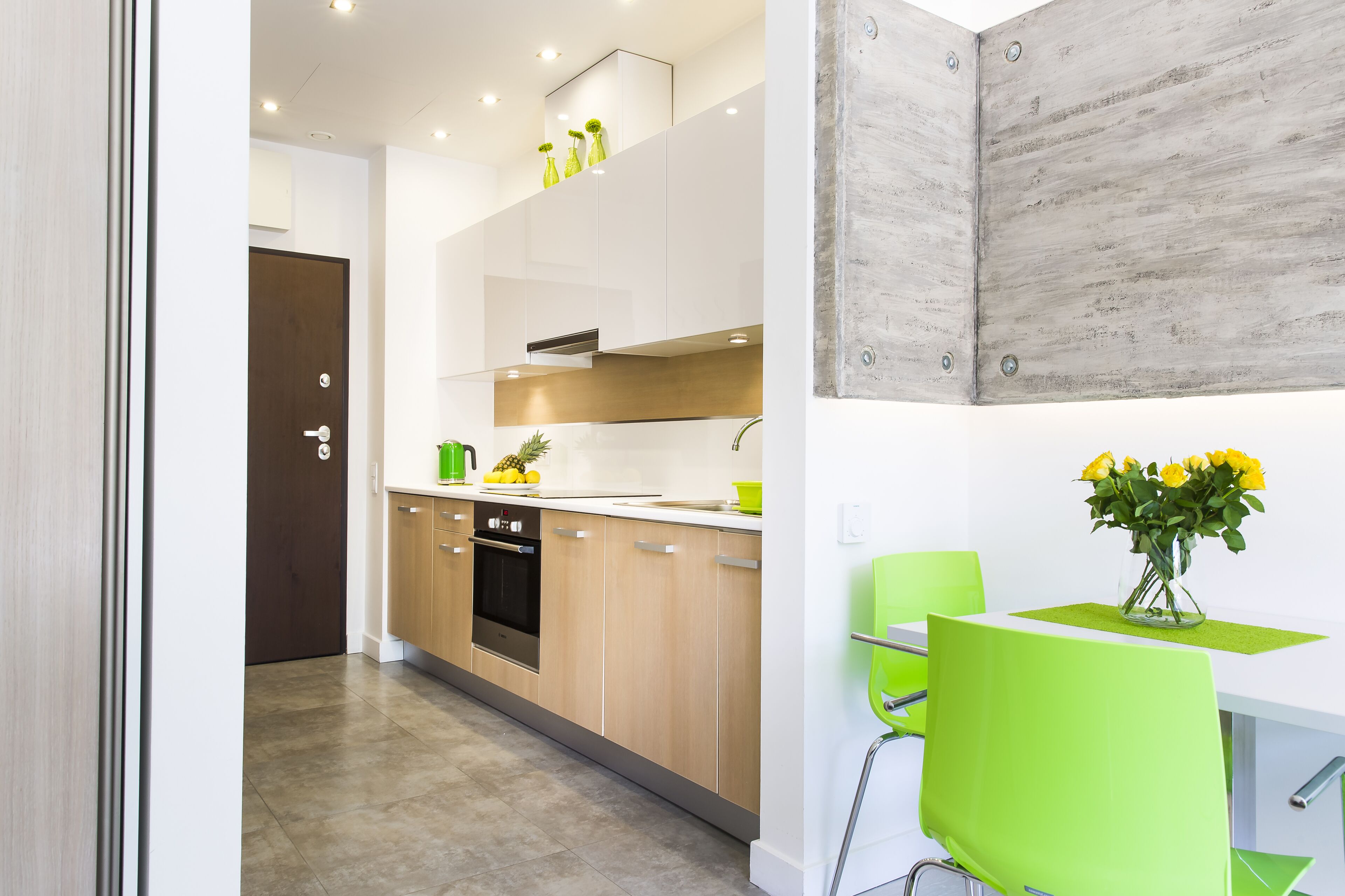A modern kitchen with wooden cabinets, a stainless steel oven, and a dining area with a green chair and a vase of yellow flowers.
