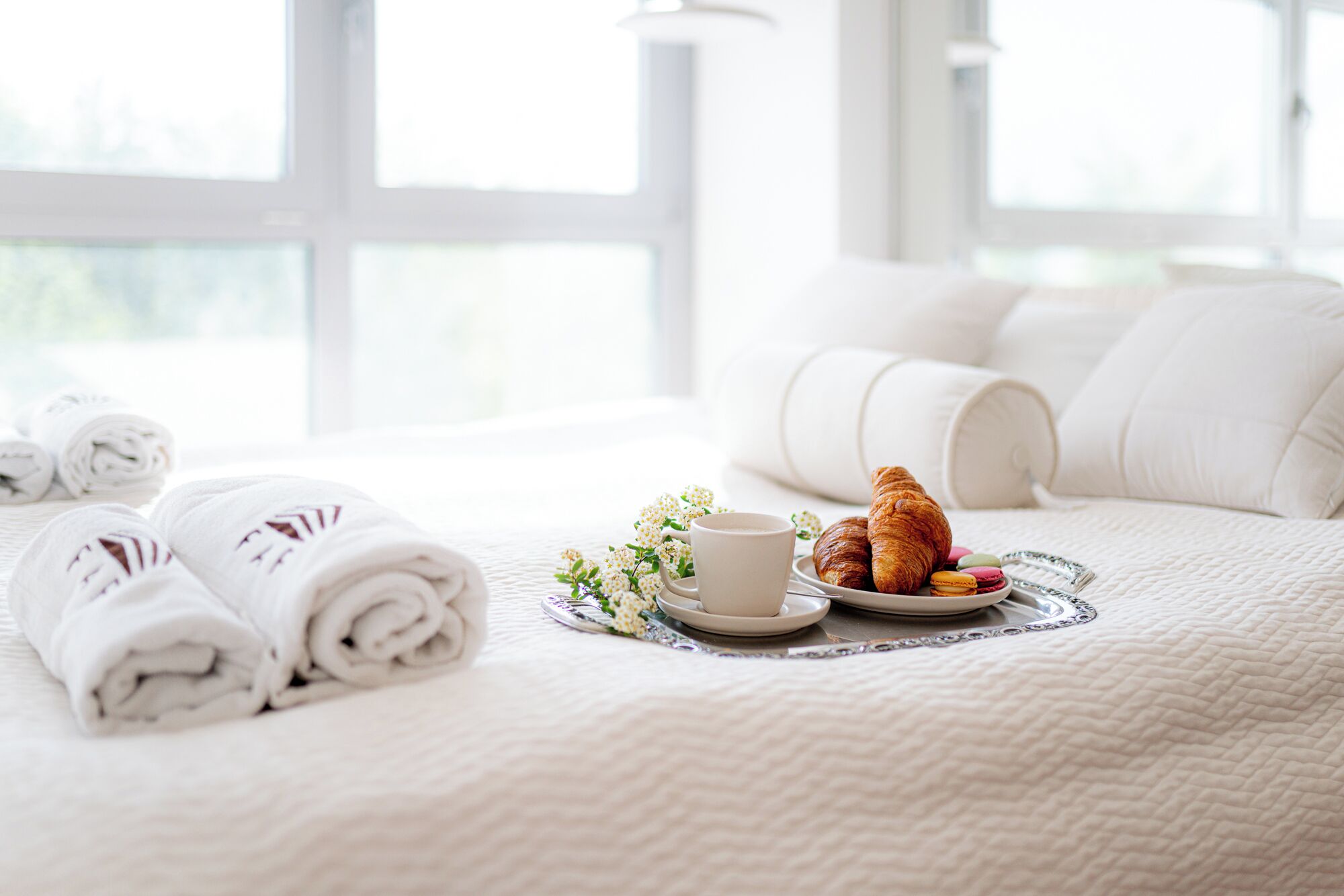 A neatly made bed with rolled white towels, a plate of croissants, a cup of coffee, and a small bouquet of flowers.