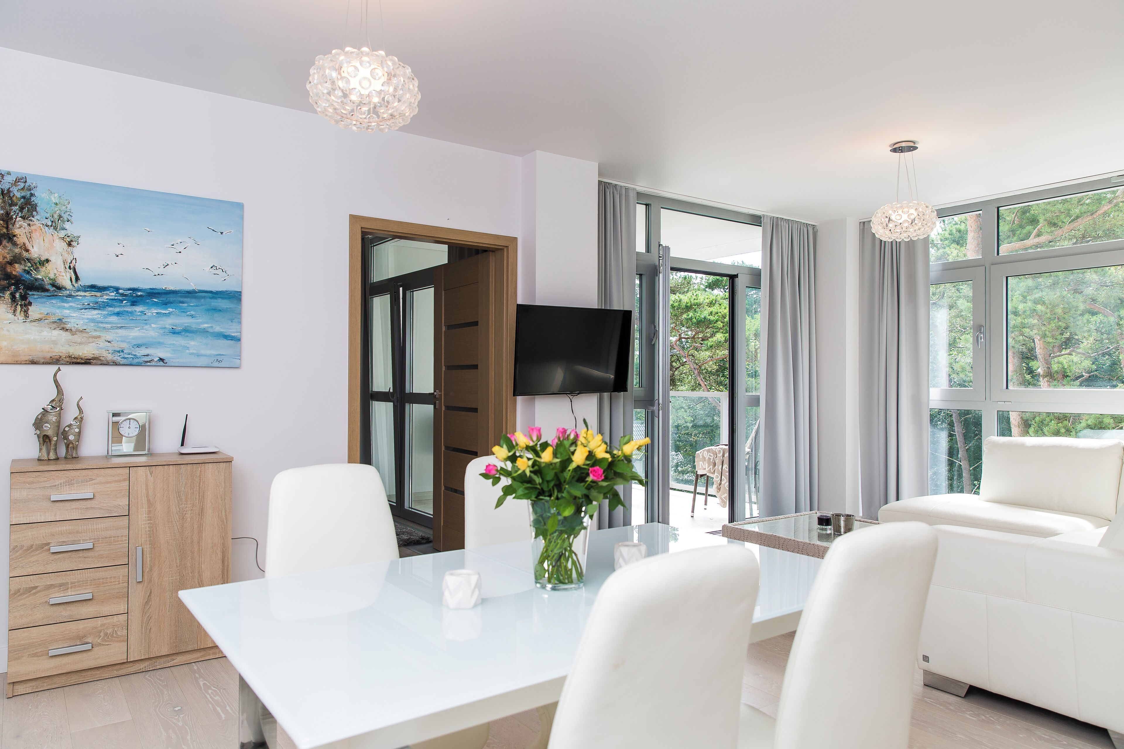A modern living room with a white sofa, a dining table with chairs, a TV, and a painting of a beach scene.