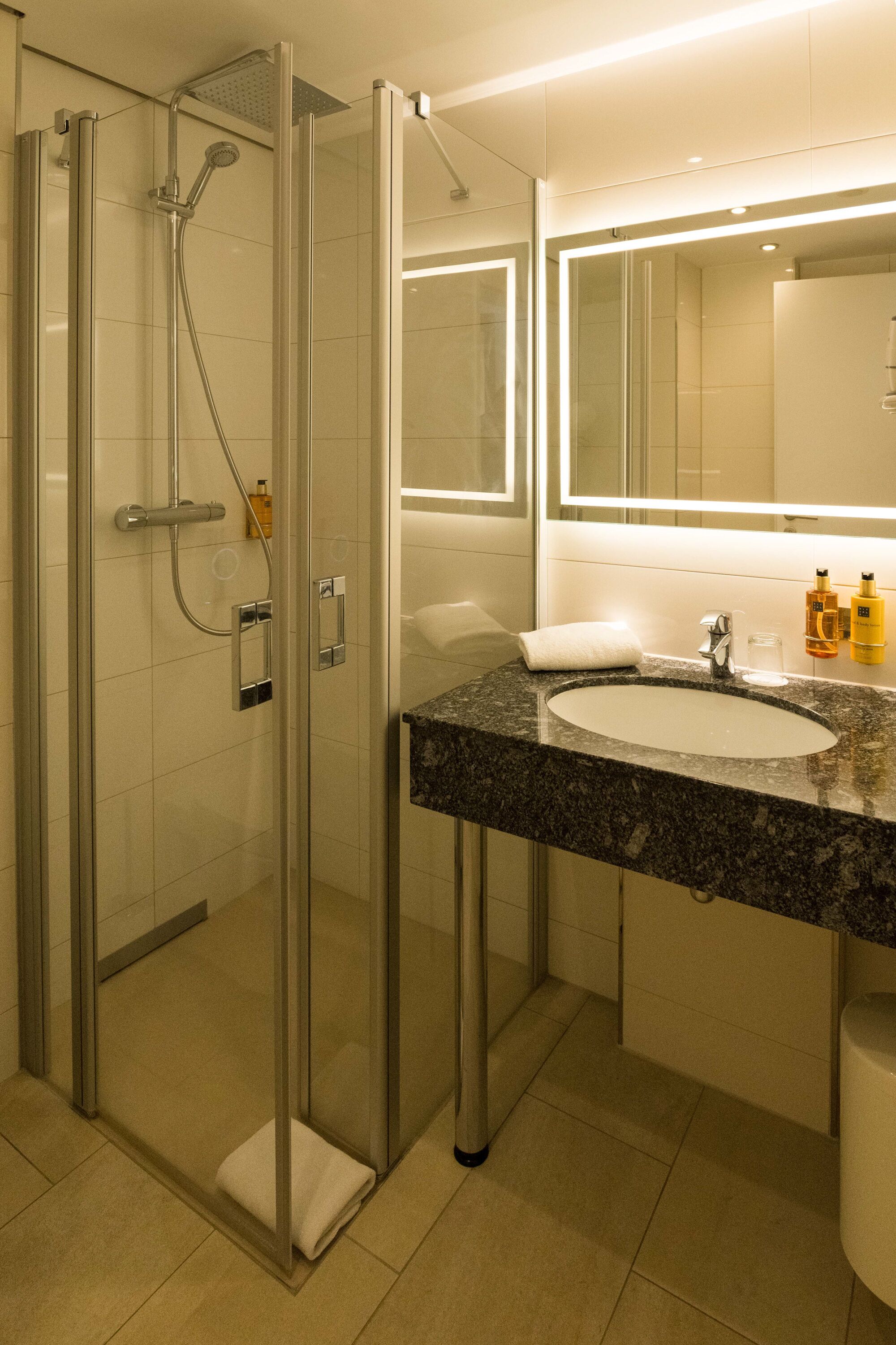 Un baño moderno con ducha envuelta en vidrio, lavamanos con encimera de granito y un espejo grande sobre el lavamanos.