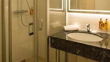 Un baño moderno con ducha envuelta en vidrio, lavamanos con encimera de granito y un espejo grande sobre el lavamanos.