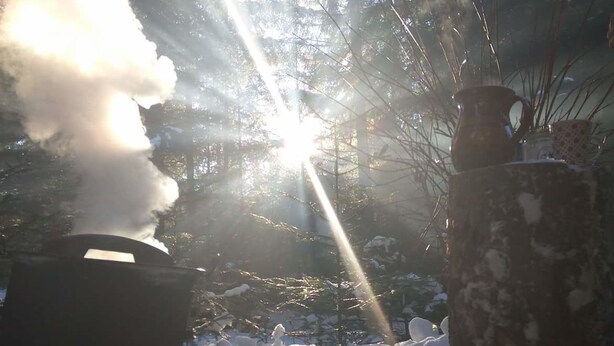 Una fogata con humo ascendente, rodeada de nieve y árboles, con la luz del sol filtrándose entre ellos.