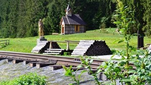 Una pequeña iglesia de madera con un campanario, rodeada de zona boscosa y vías de tren.