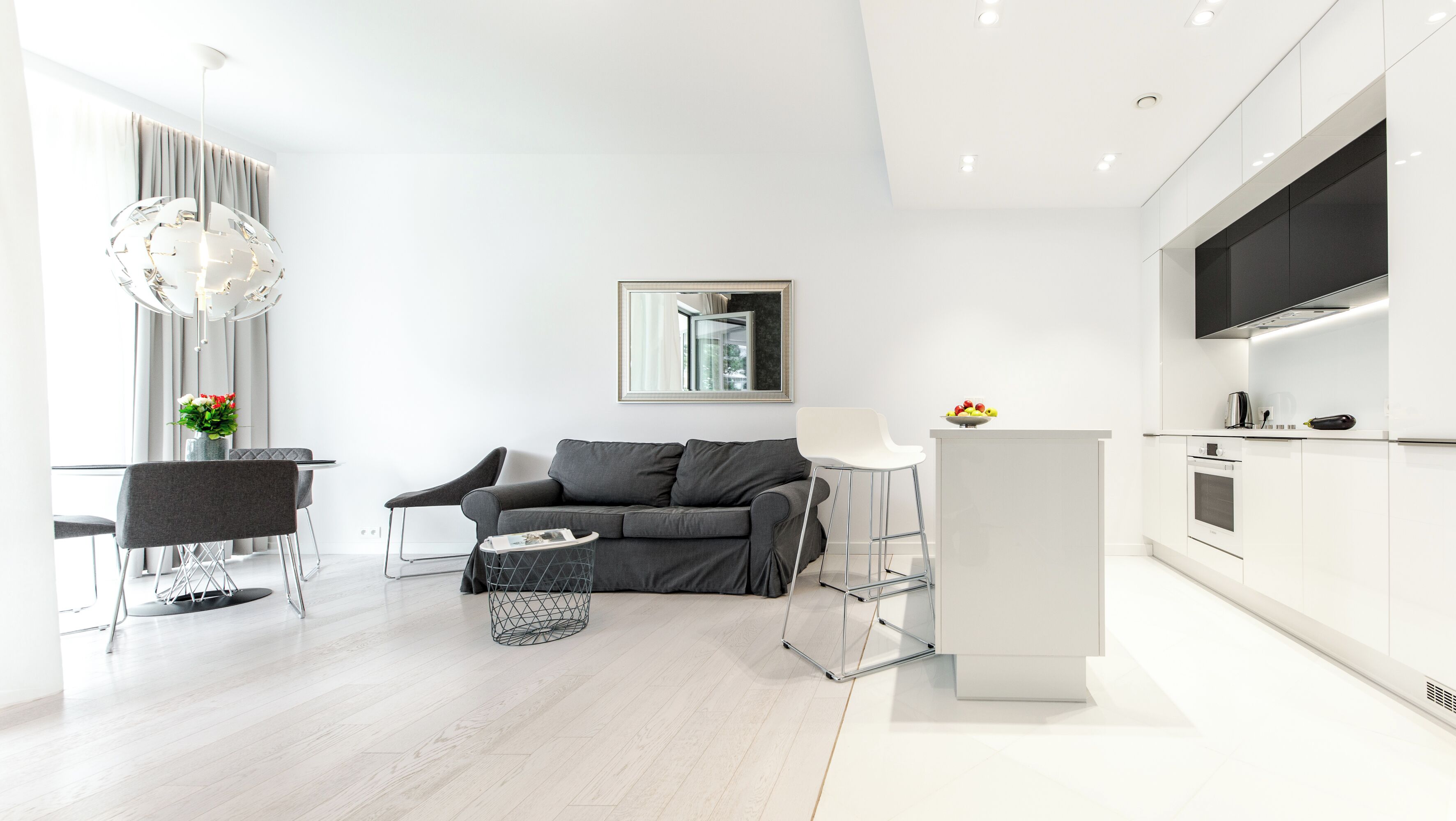 Uma sala de estar moderna e minimalista com um sofá cinza, uma mesa de centro e um espaço de jantar com uma mesa e cadeiras brancas. Uma cozinha com armários e eletrodomésticos brancos é visível à direita.