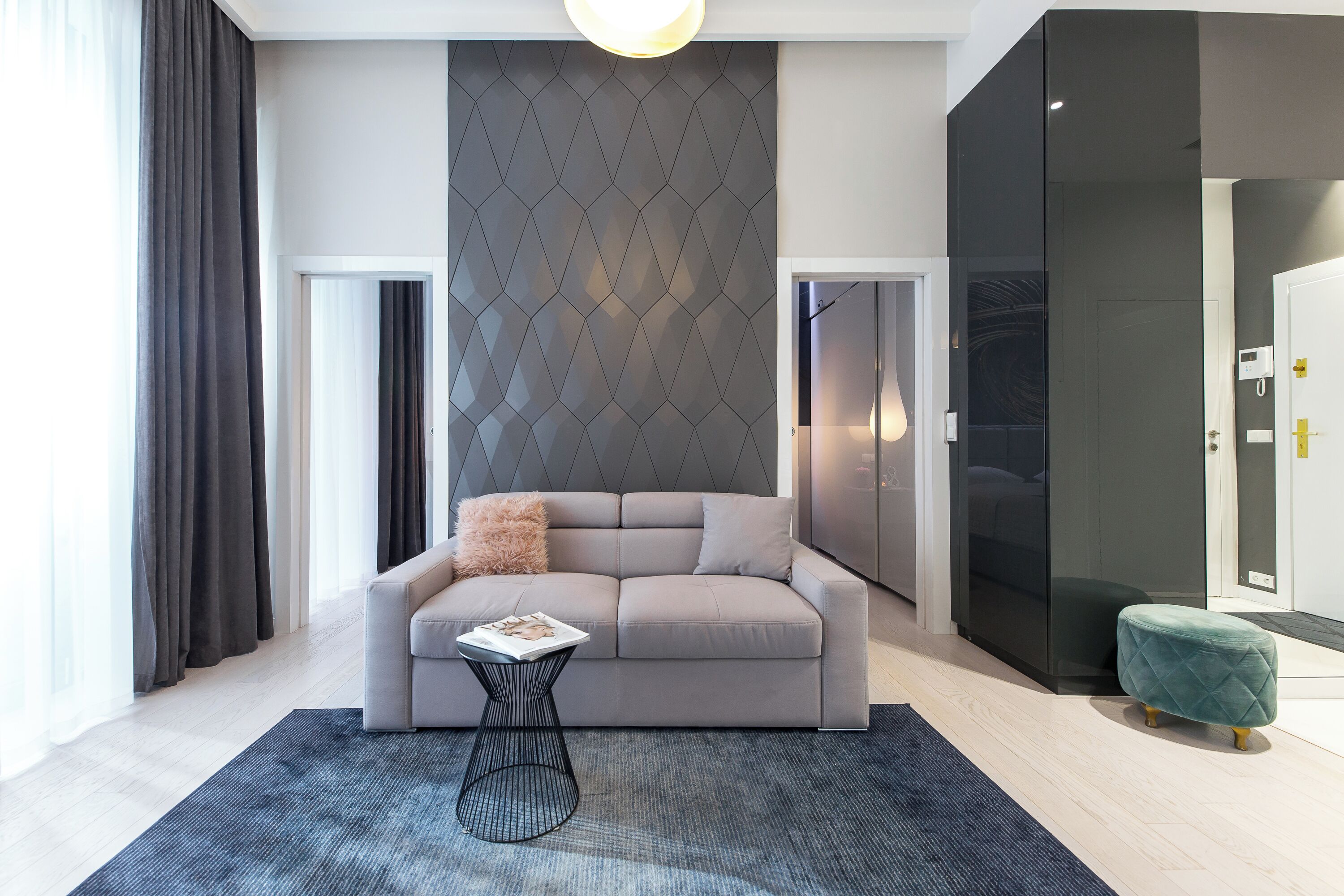 Un salón moderno con un sofá gris, una mesita auxiliar, un papel tapiz estampado y un amplio ventanal con cortinas.
