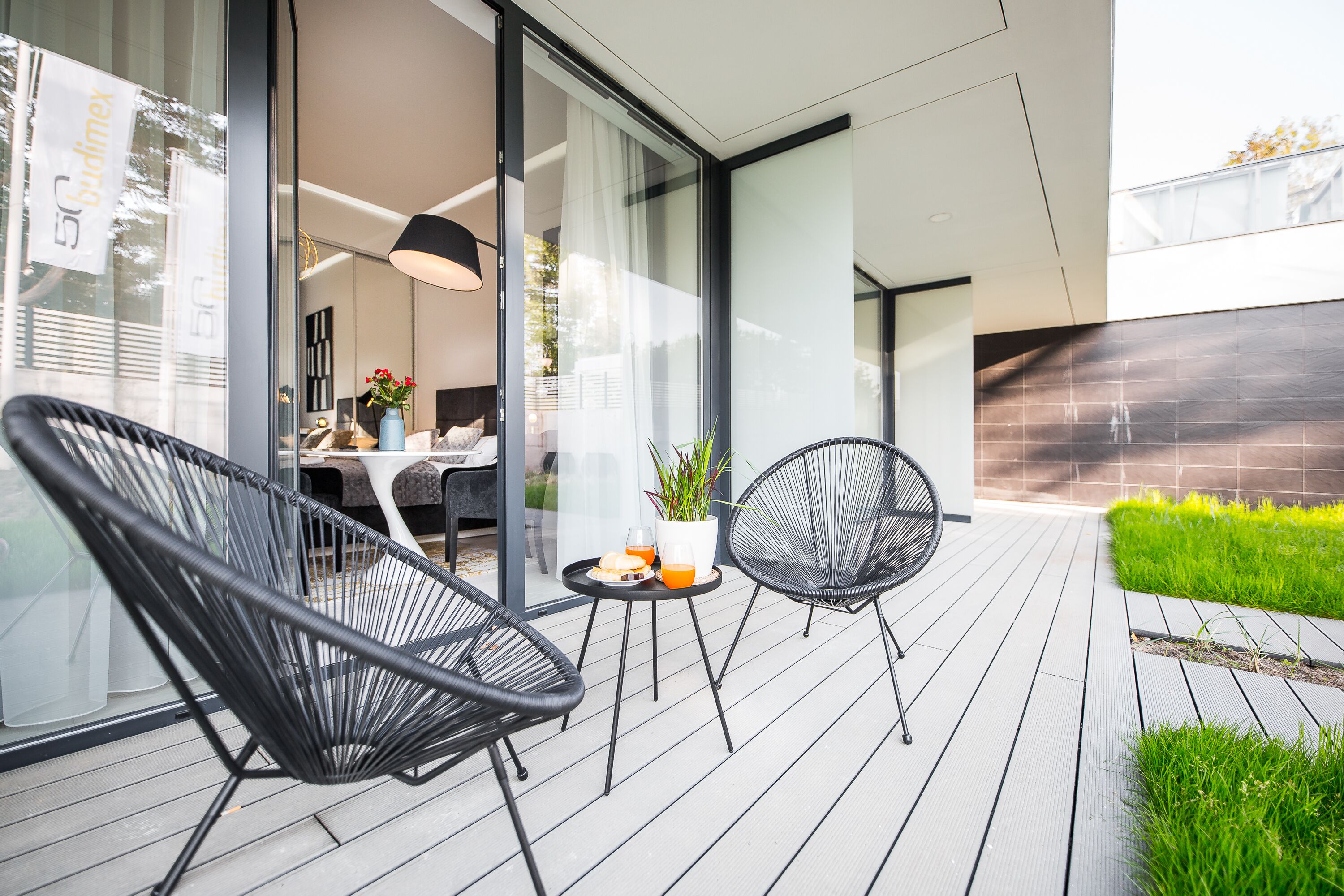 A modern outdoor patio with two black wireframe chairs, a small table with a plant and a glass of orange juice, and a glass door leading to an indoor area.