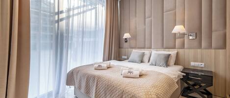 A modern bedroom with a large bed, a bedside table with a lamp, and a view through sheer curtains.