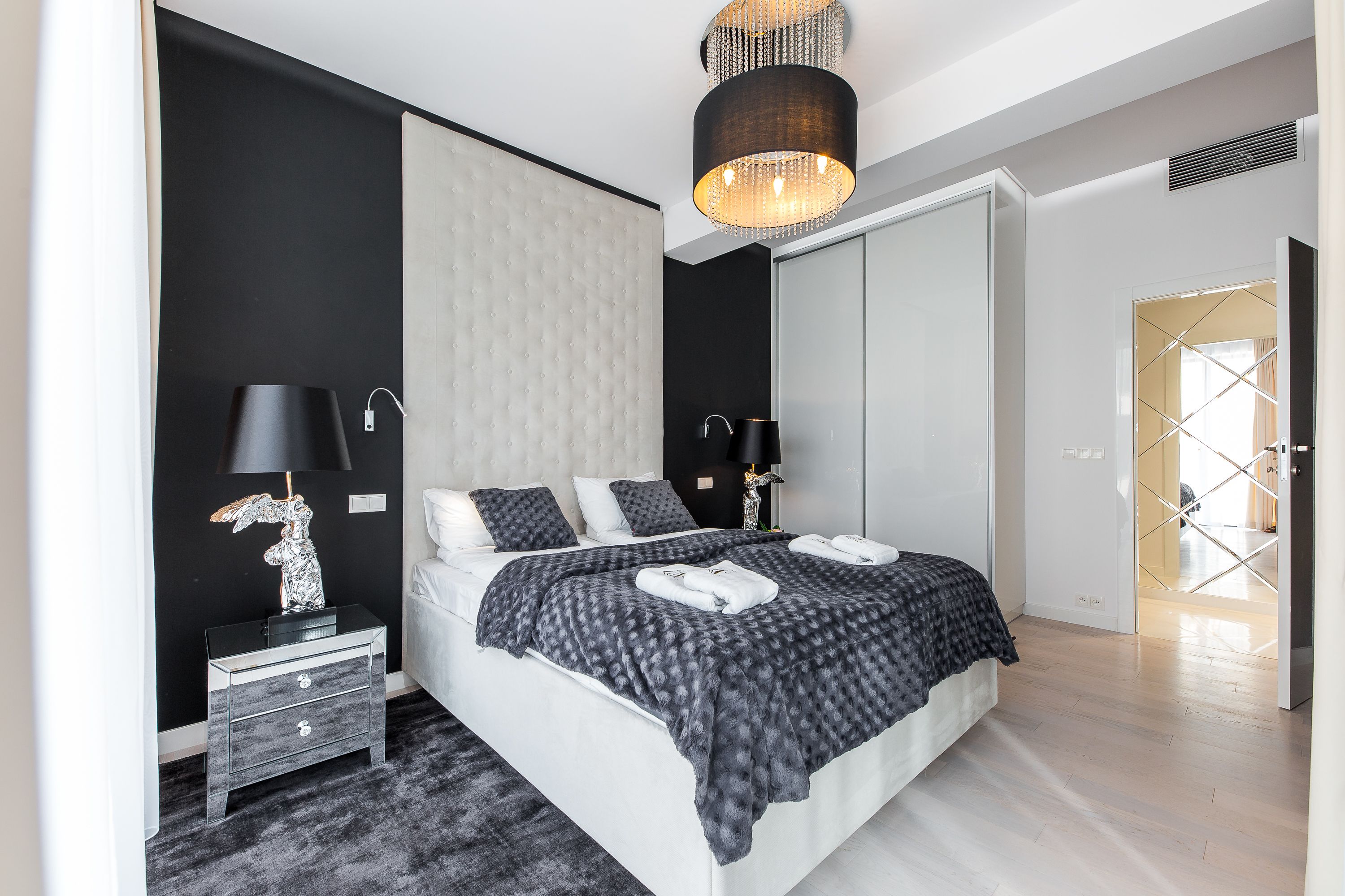 A modern bedroom with a large bed, a nightstand, a lamp, and a mirror.