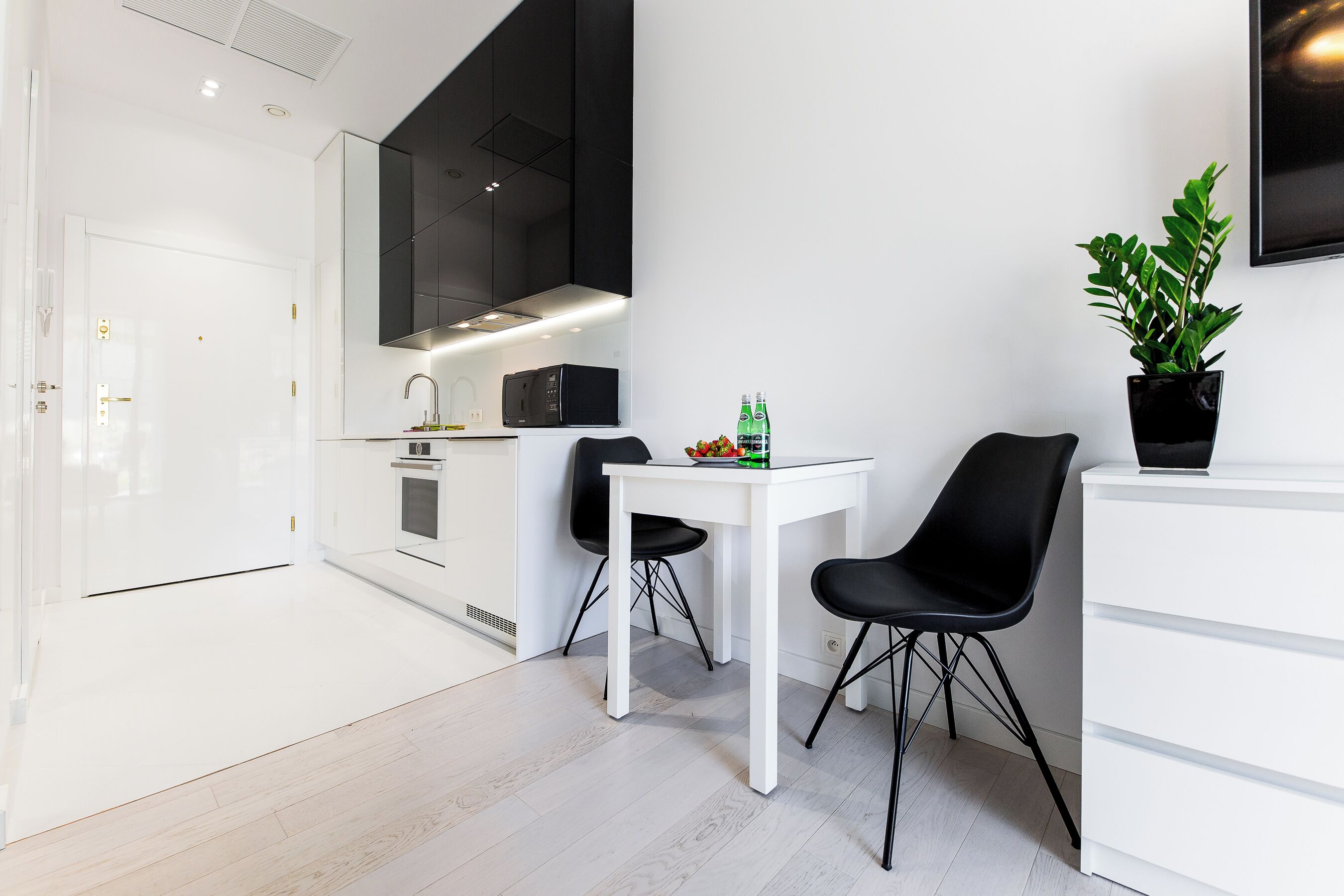 A modern kitchen with white cabinetry, a black microwave, and a white dining table with two black chairs. There is a potted plant on a white cabinet and a bottle of water on the table.