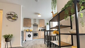 A modern kitchen with wooden cabinets, a built-in washing machine, and a dining area with a black metal shelf unit. - Grand Central Littleover Deluxe (Derby)