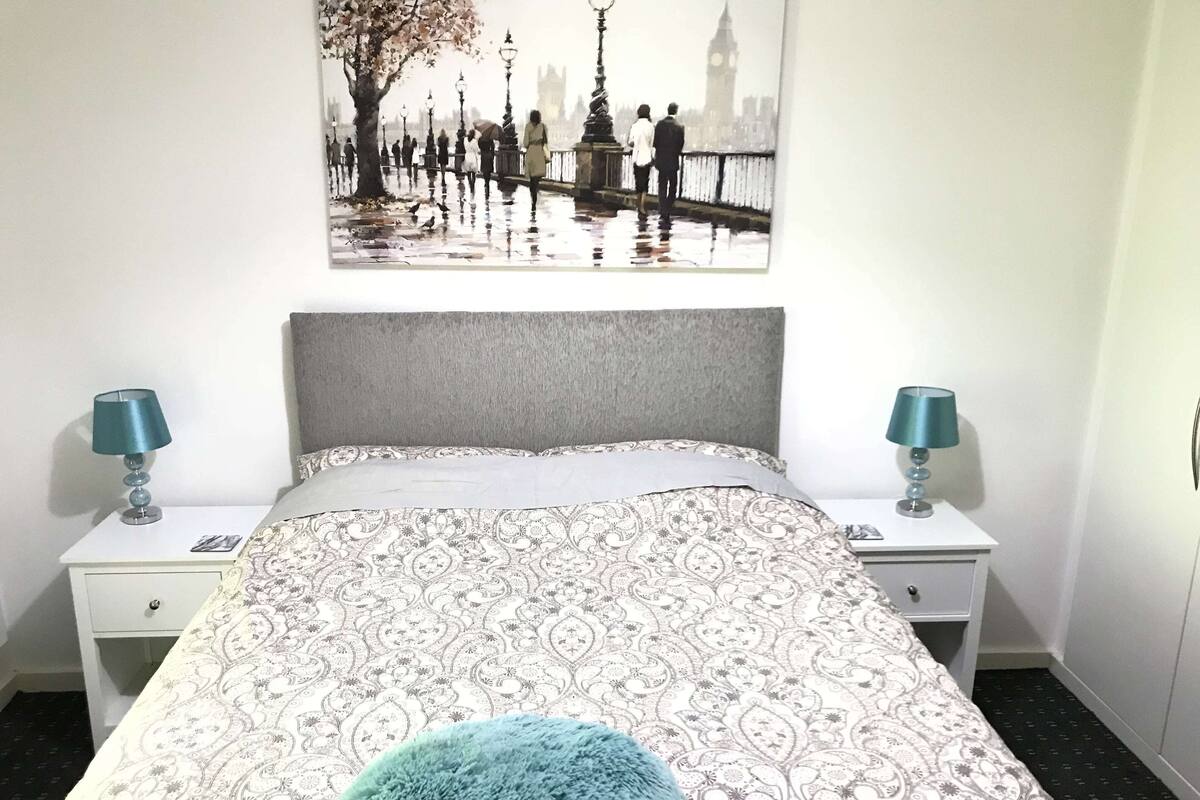A bedroom with a patterned bedspread, a grey headboard, two bedside tables with lamps, and a framed picture of a cityscape.