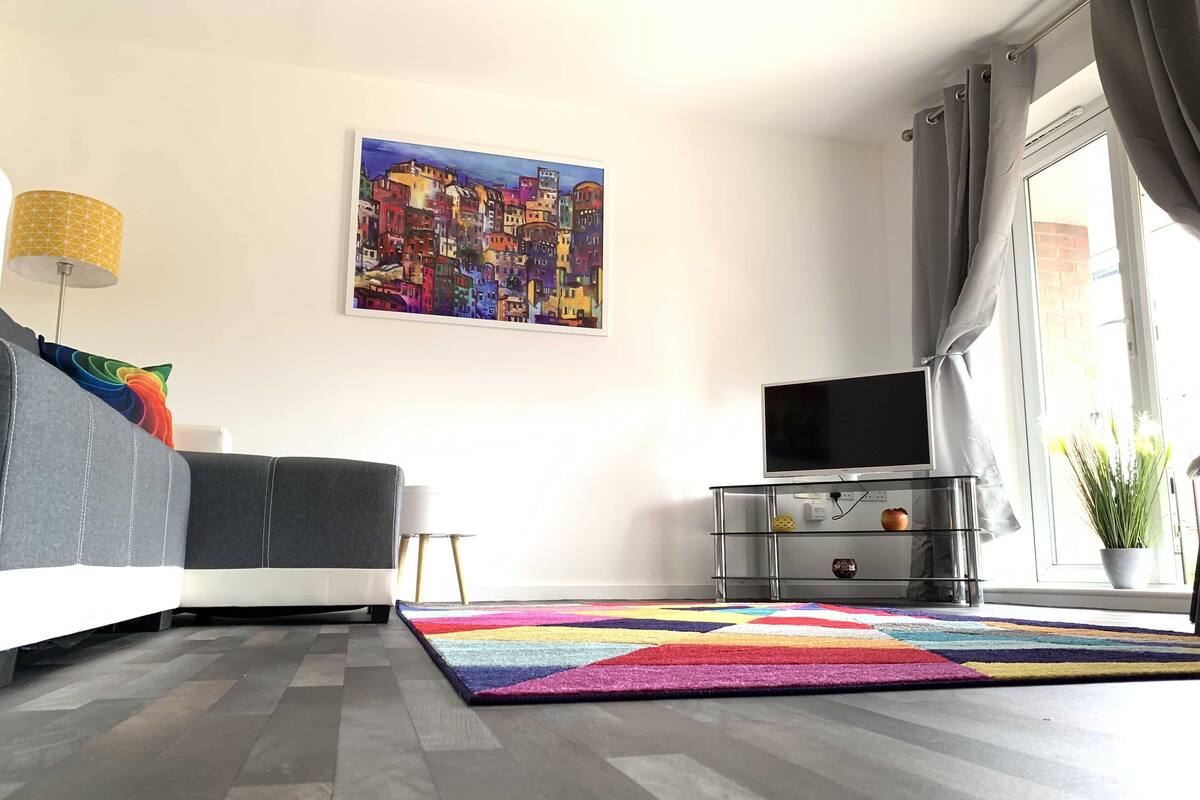 A modern living room with a grey sofa, a colorful rug, a flat-screen TV on a glass cabinet, and a cityscape painting on the wall.
