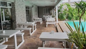 An outdoor patio area with white tables and benches, a stone wall, and a pool. - Savoy hotel (Jeffreys Bay)