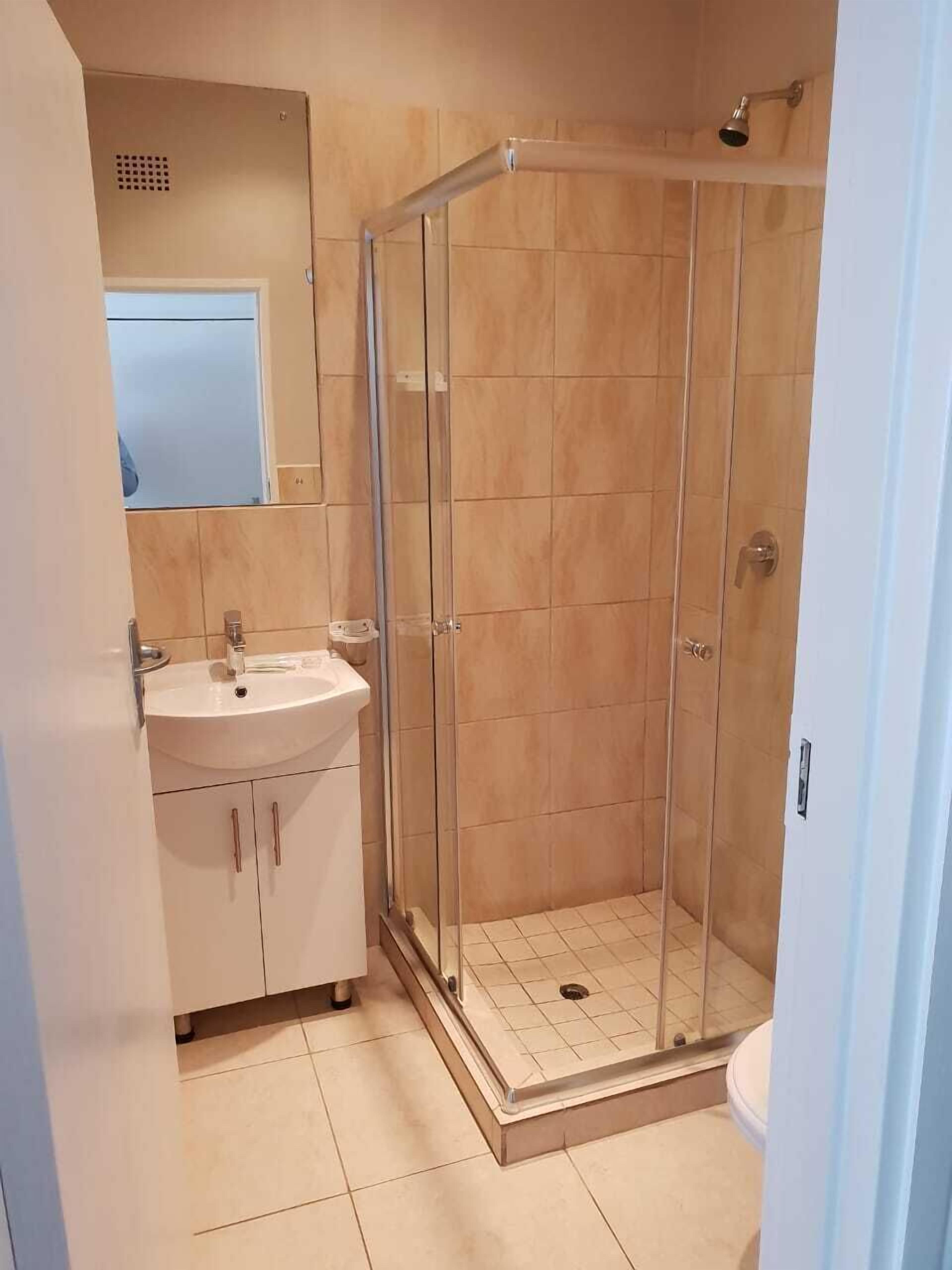 Un baño con ducha envuelta en cristal, lavamanos con espejo y paredes de baldosas color beige.