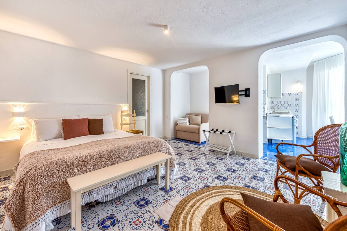 Habitación de hotel con una cama, un banco, una silla de mimbre, un televisor y un baño visible a través de un arco.