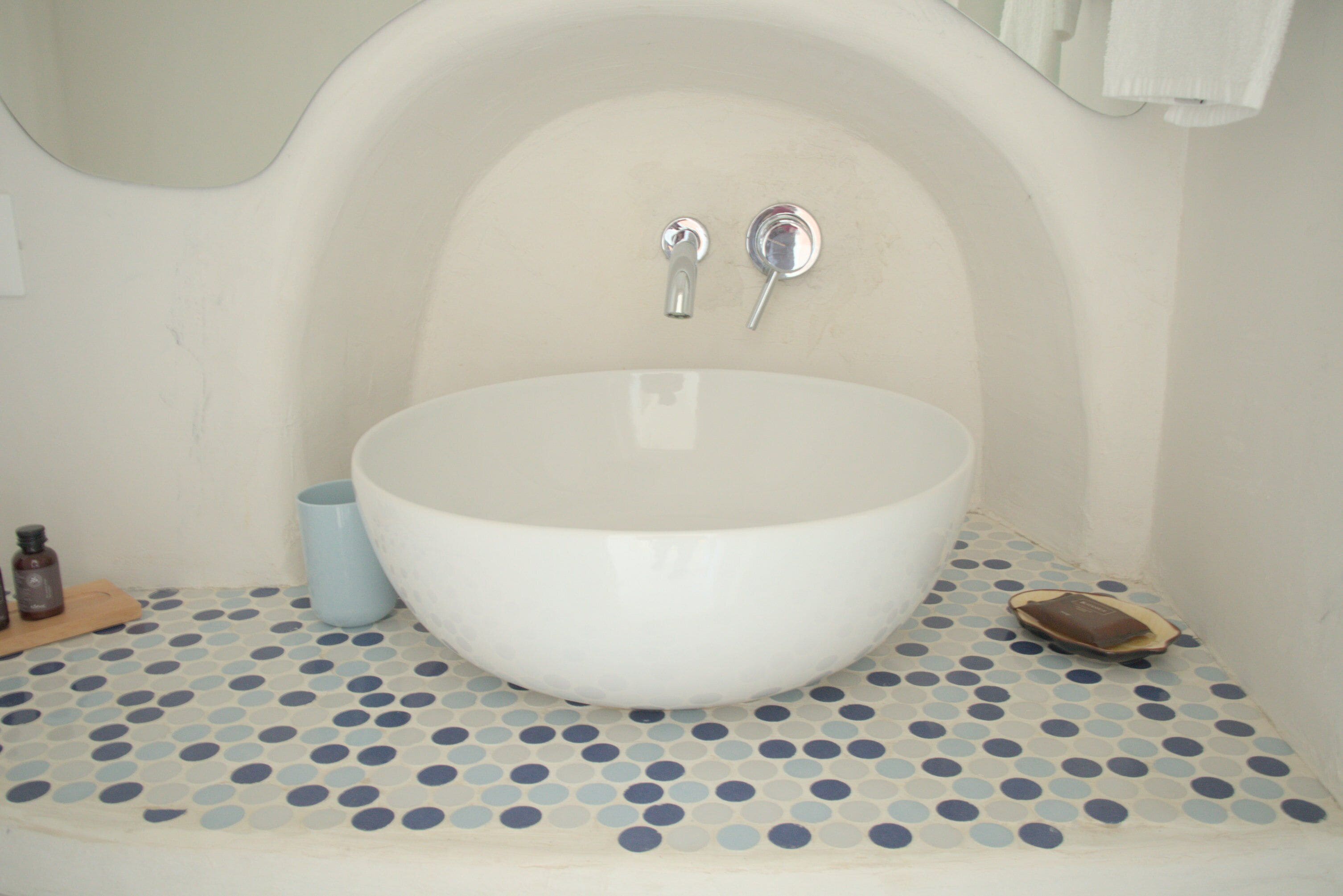 Un baño con un lavamanos grande de color blanco, piso azul y blanco a lunares, y un estante de madera pequeño con artículos de aseo.