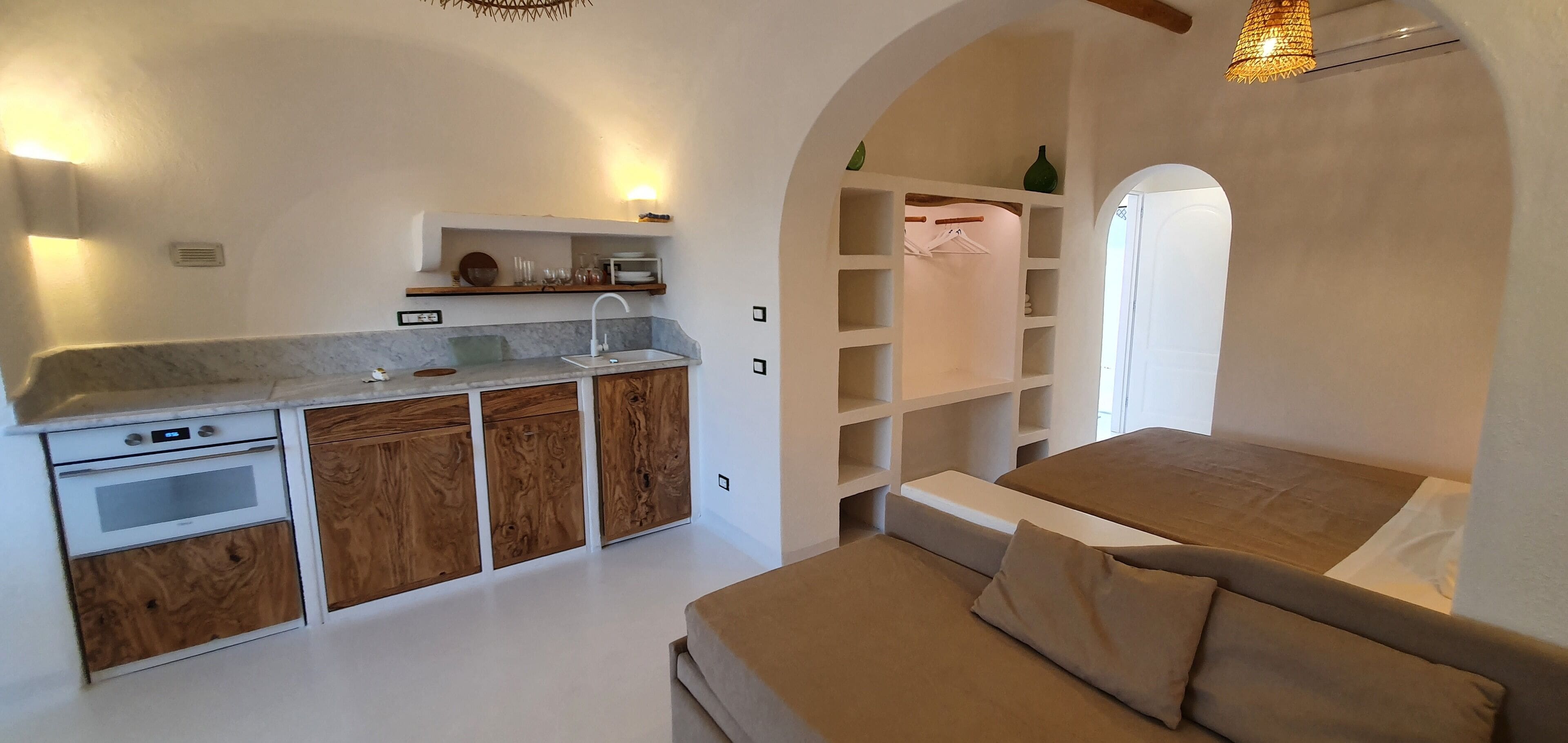 A compact bedroom with a built-in kitchenette, a bed with a headboard, and a wardrobe with hanging space.