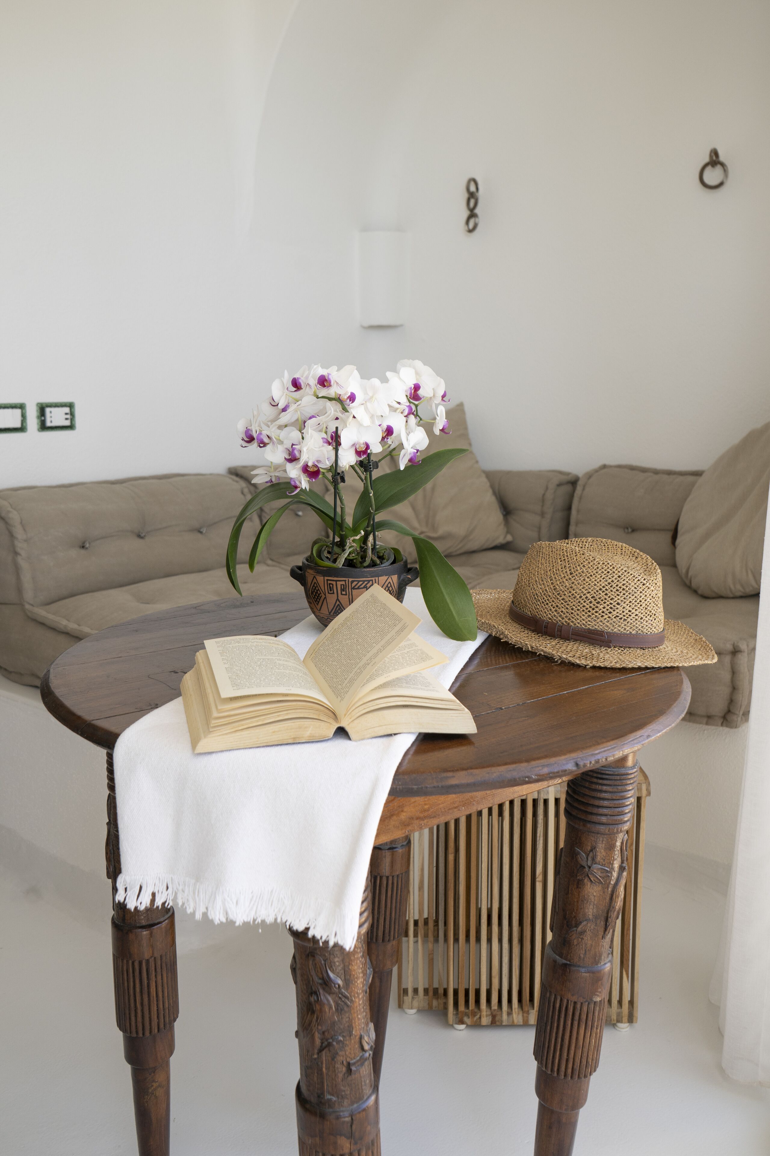 Une table ronde en bois, sur laquelle se trouvent un chapeau tressé et un livre ouvert, un pot d’orchidée et, au fond, un canapé beige.