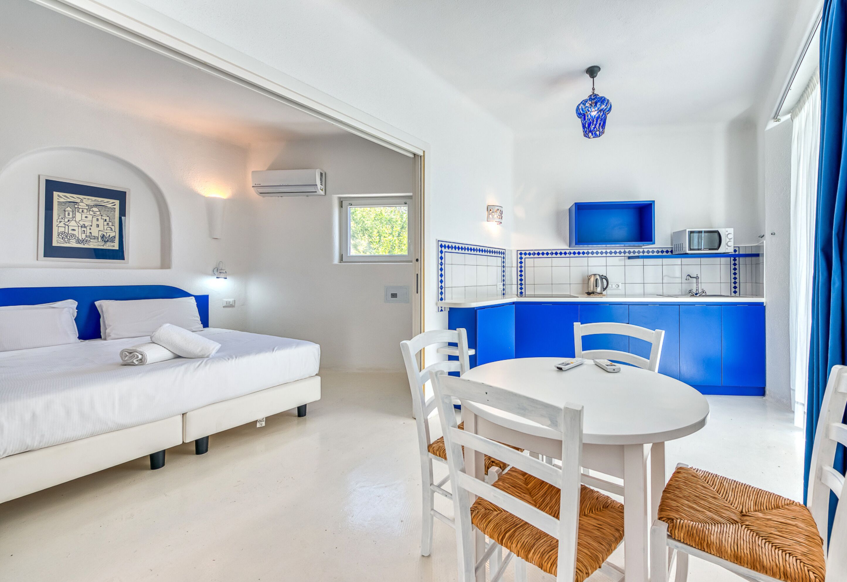 A bedroom with a bed, a bedside table, and a window. There is a kitchenette with a microwave and a sink.