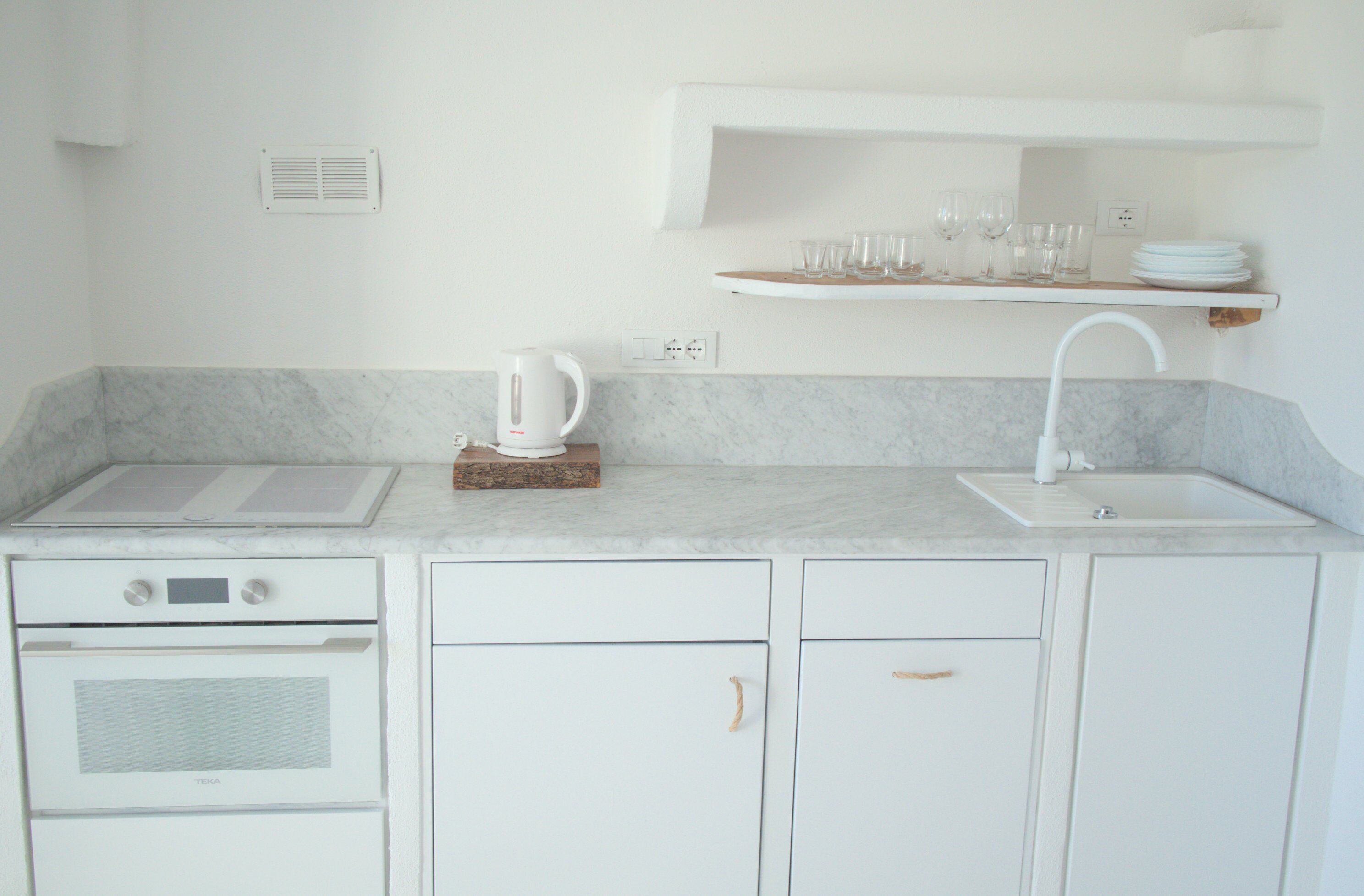 A kitchen with white cabinets, a built-in oven, a sink, and a shelf above the counter holding glasses and plates.