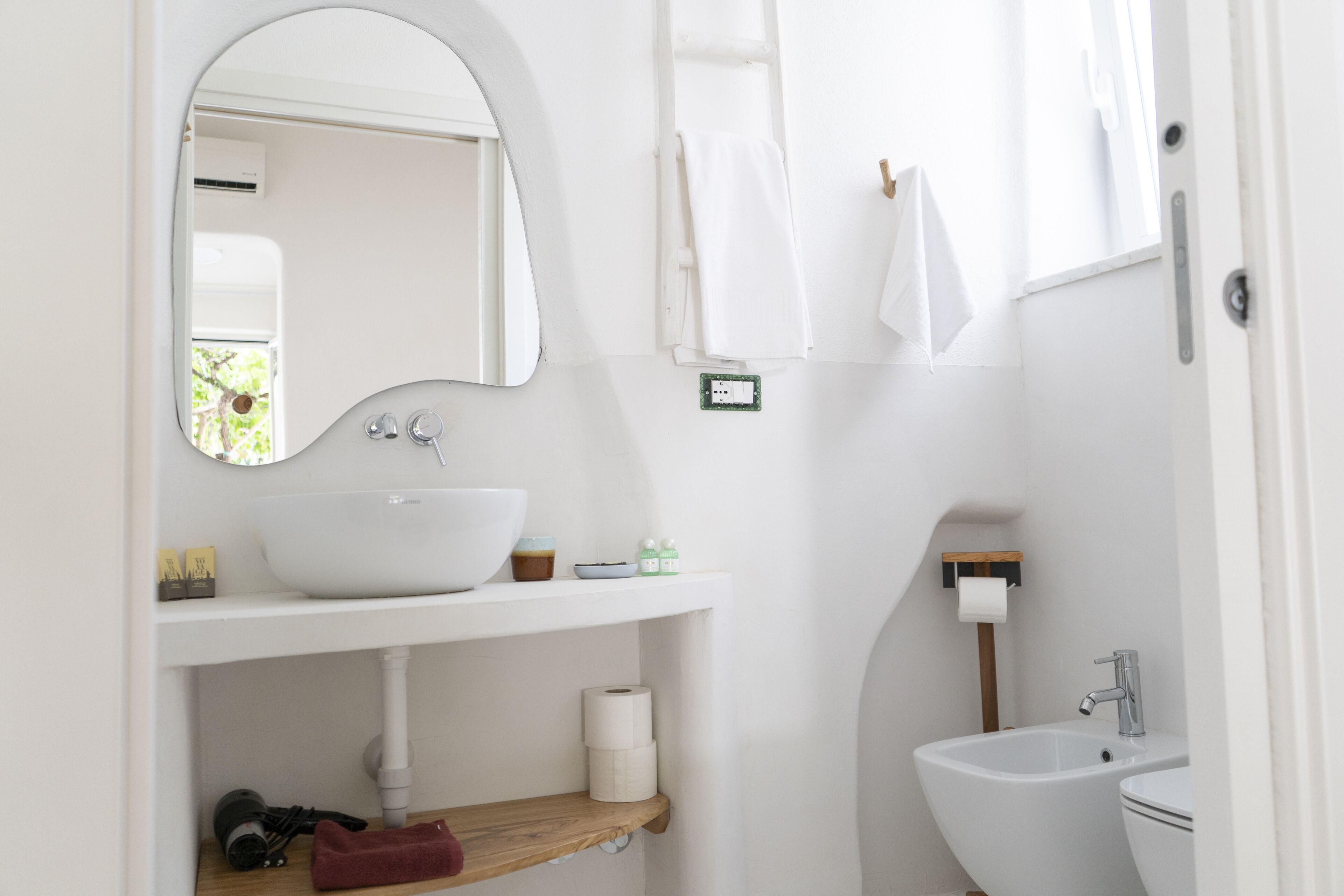 Un baño con lavamanos blanco, espejo y retrete. Hay un perchero, un estante con artículos de aseo y una ventana con cortinas.