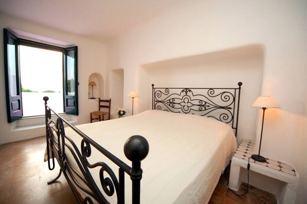 Un dormitorio con cama, ventana con vistas, mesita de noche con lámpara y una silla.