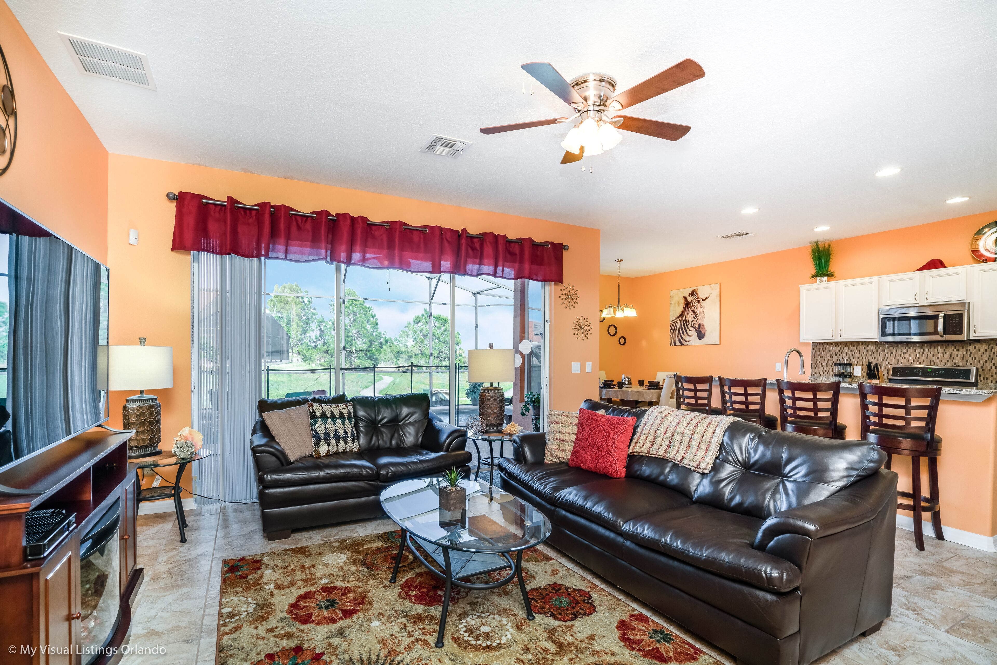 A living room with two leather sofas, a glass coffee table, and a red rug. There is a ceiling fan, a kitchen area with white cabinets, and a dining area with wooden chairs.