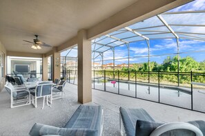 Un porche cubierto con mesa y sillas, ventilador de techo y vistas a una piscina y follaje.