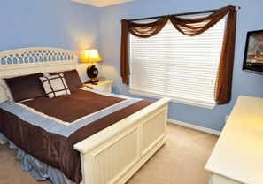 Un dormitorio con cama, mesita de noche, lámpara, ventana con cortinas e imagen colgada en la pared.