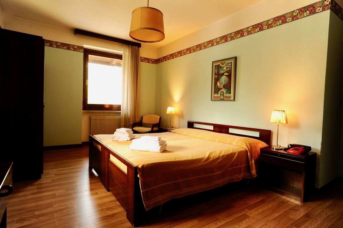 Ein Hotelzimmer mit einem großen Bett, zwei Nachttischlampen, einem Holznachttisch mit Telefon, einem Sessel, einem Fenster mit Vorhängen und einer floralen Bordüre an den Wänden.