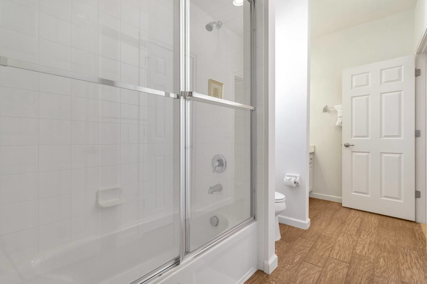 Une salle de bain moderne avec une douche à parois de verre, des murs recouverts de carreaux blancs, des toilettes et un plancher en bois.