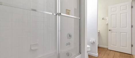 Un baño moderno con ducha de vidrio, paredes cubiertas de azulejos blancos, inodoro y suelo de madera.