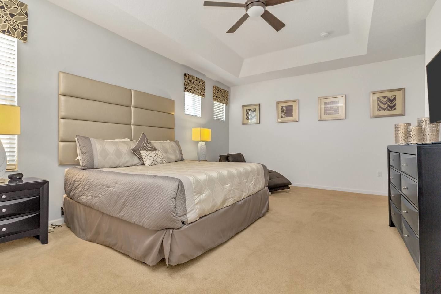 A bedroom with a large bed, a nightstand, a dresser, a ceiling fan, and framed pictures on the wall.
