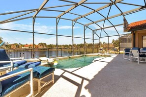 Piscina exterior techada con sillas de descanso y una mesa, rodeada por una valla metálica y vistas a un cuerpo de agua y árboles.