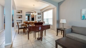 A spacious living room with a piano, a sofa, and a dining area with chairs and a table. - Emerald Island - 7bd/4.5ba Pool Home - #7ei565 (Kissimmee)