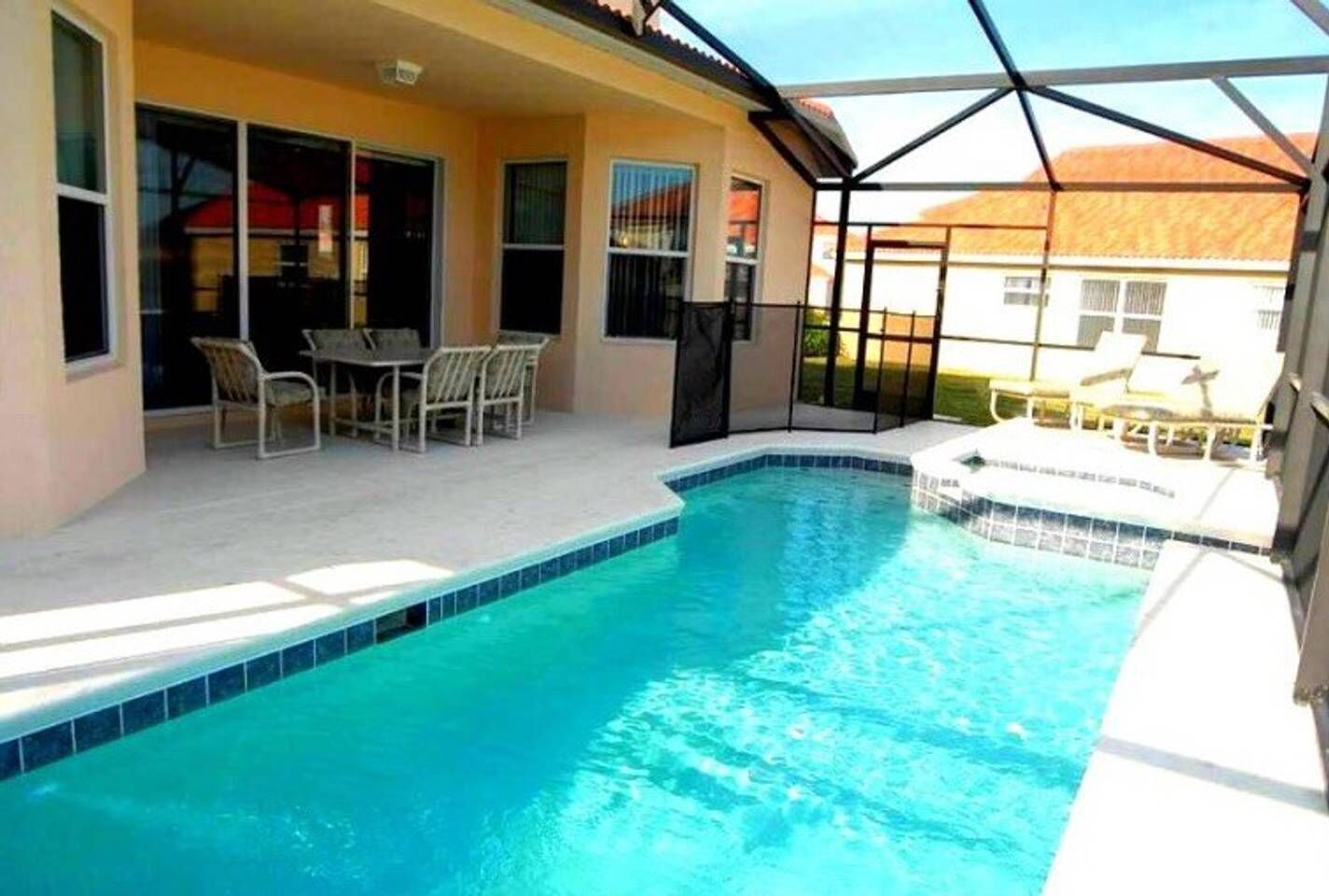 A swimming pool with a sunshade structure, a patio with chairs, and a view of a residential area.