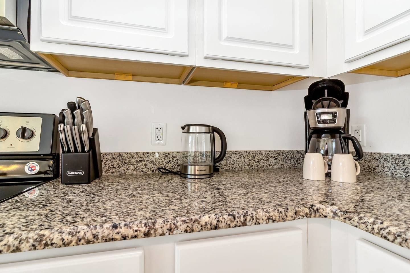 Una cocina con armarios blancos, encimera de granito, estufa, cafetera y hervidor.