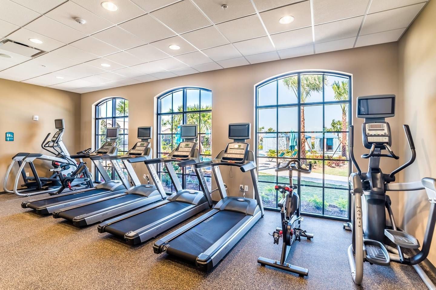 Een sportschool met loopbanden, elliptische trainers en een hometrainer, grote ramen met uitzicht op groen, en een plafond met inbouwverlichting.