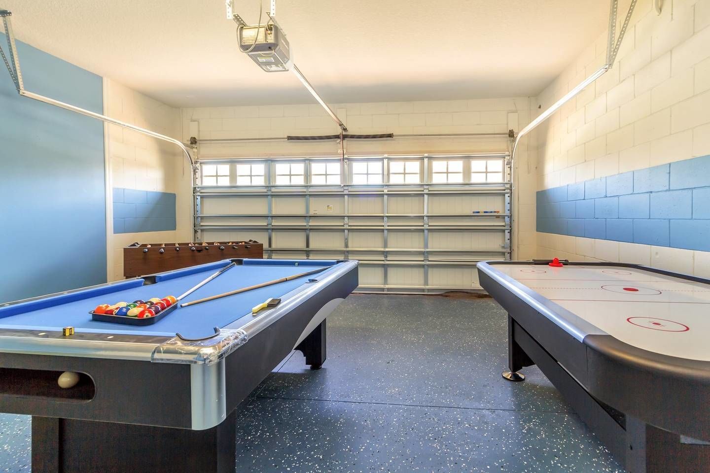 Une table de billard avec des billes et des queues, une table de baby-foot, et une grande fenêtre avec des stores.