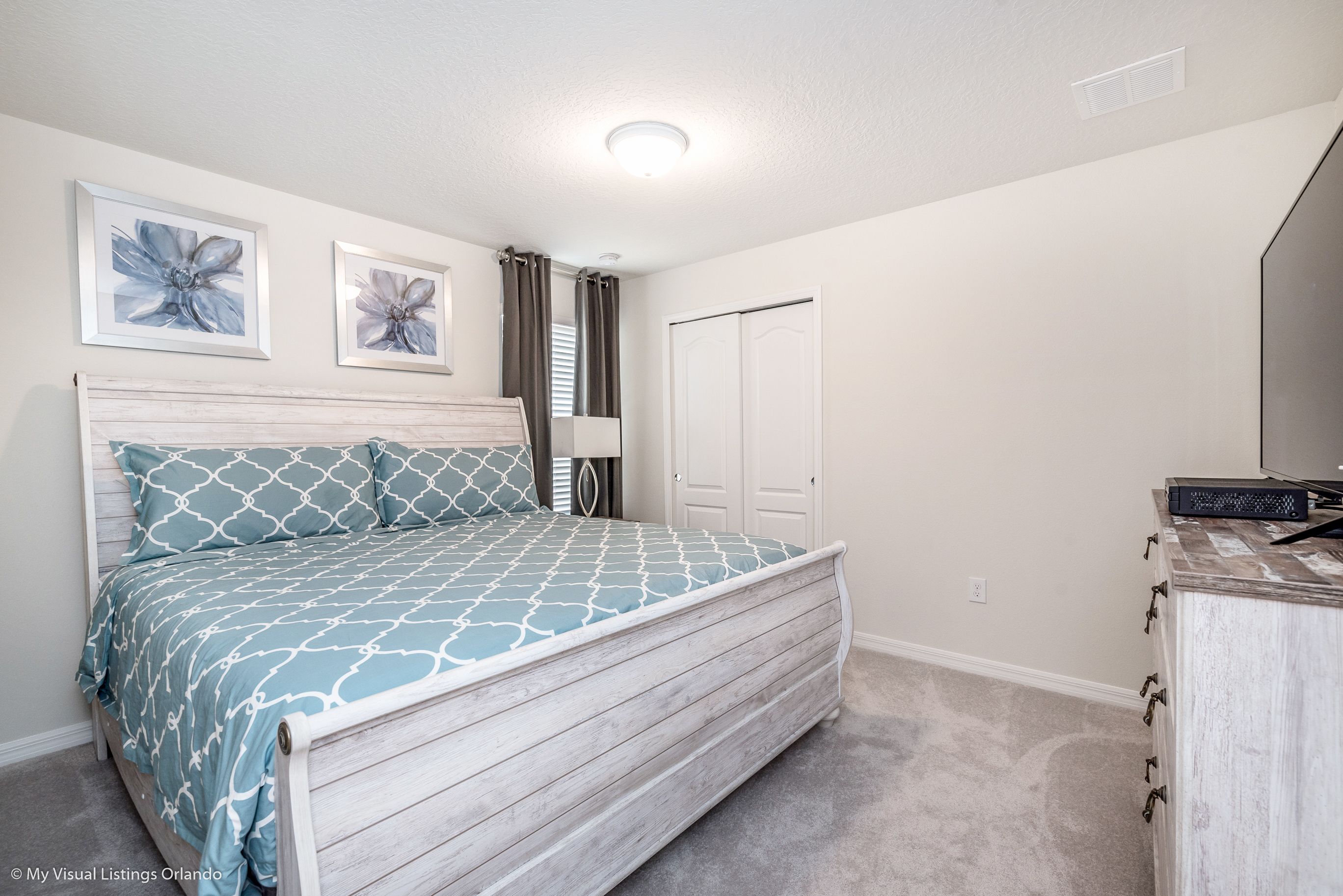 A bedroom with a bed, two framed floral wall art pieces, a white door, and a dresser with drawers.