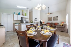 A dining area with a wooden table set for six, surrounded by chairs. There is a kitchen in the background with white cabinets and a refrigerator.