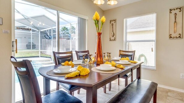 A dining area with a table set for four, a vase with yellow flowers, and a view of a backyard through large sliding glass doors. - Hampton Lakes - Spacious 4bd/2ba Pool Home #4hl510 (Davenport)
