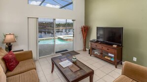 A spacious living room with a large sliding glass door leading to a pool area, a television on a wooden stand, and a lamp on a side table.