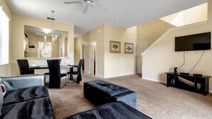 A spacious living room with a ceiling fan, a dining area with a chandelier, a staircase, and a TV mounted on the wall.
