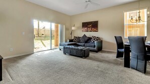 A spacious living room with a grey sofa, a matching ottoman, and a dining area with a table and chairs. There is a large window providing natural light and a view of the outdoors.